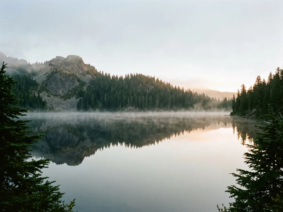 A still Cascade alpine lake at dawn mirroring the evergreen ridge above it, with thin mist on the water.